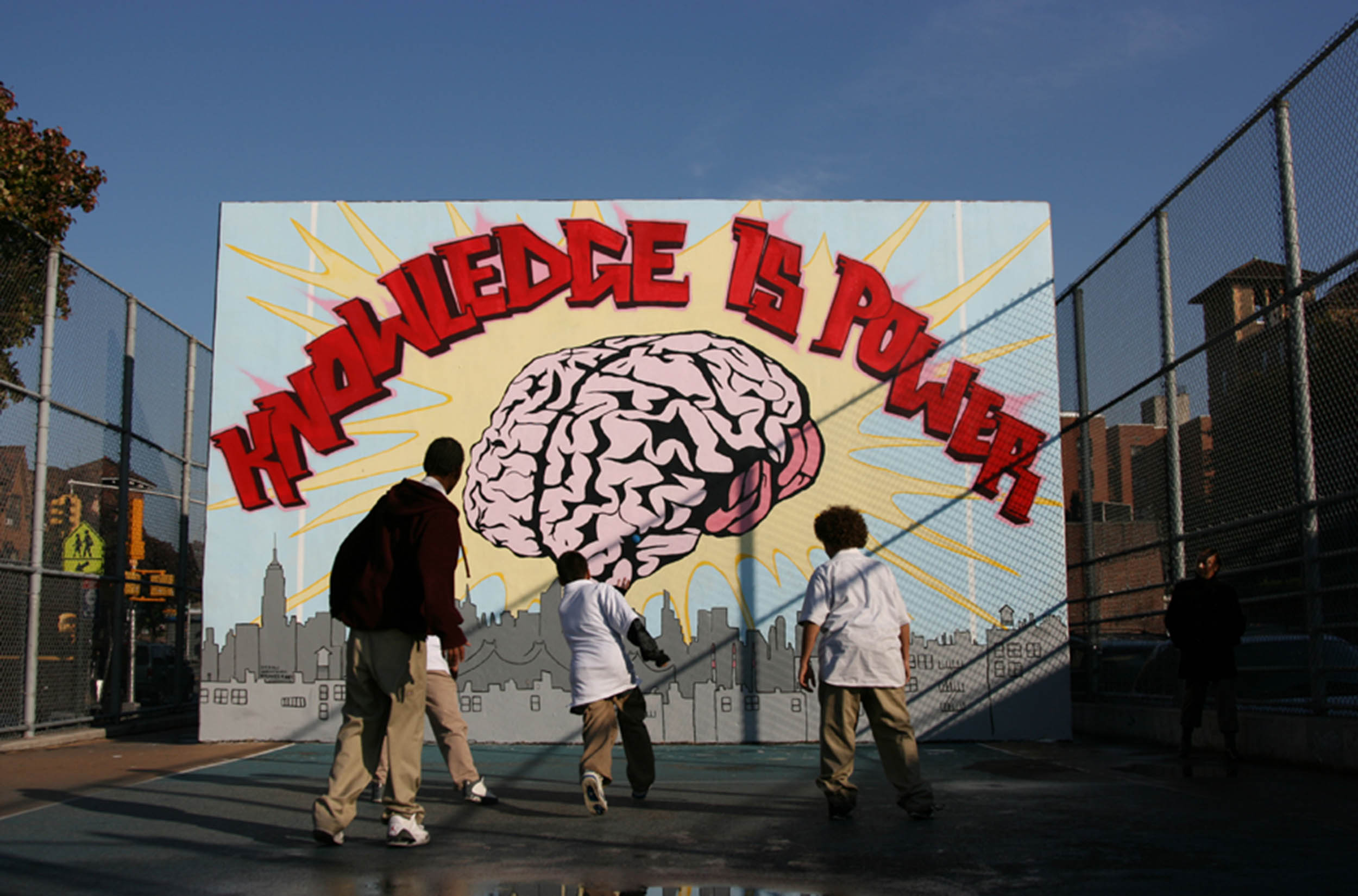 Mural on a handball wall depicting a city skyline, a photo-realistic depiction of a human brain, and the words Knowledge is Power. Three people play handball against the wall.