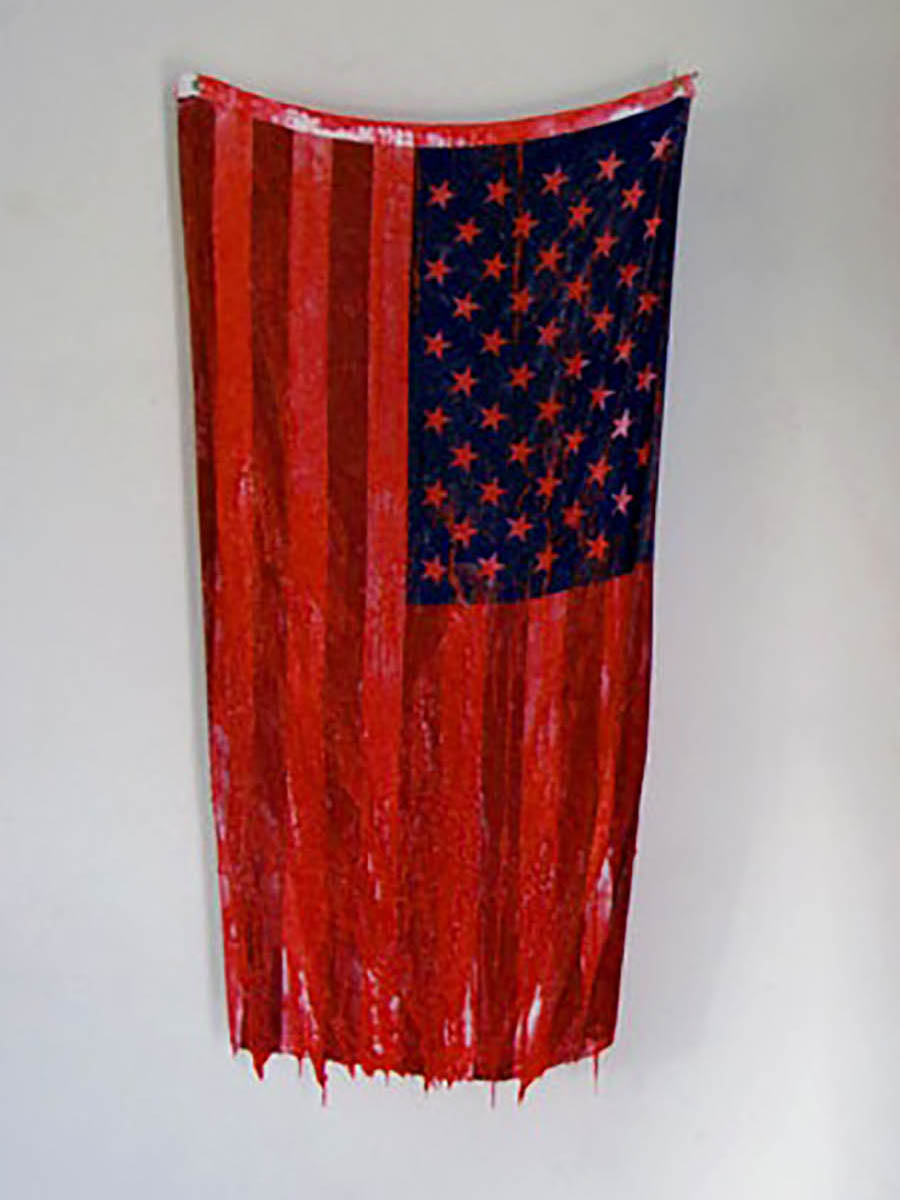 An artwork comprised of an American flag hung vertically and dipped in red paint to resemble blood.