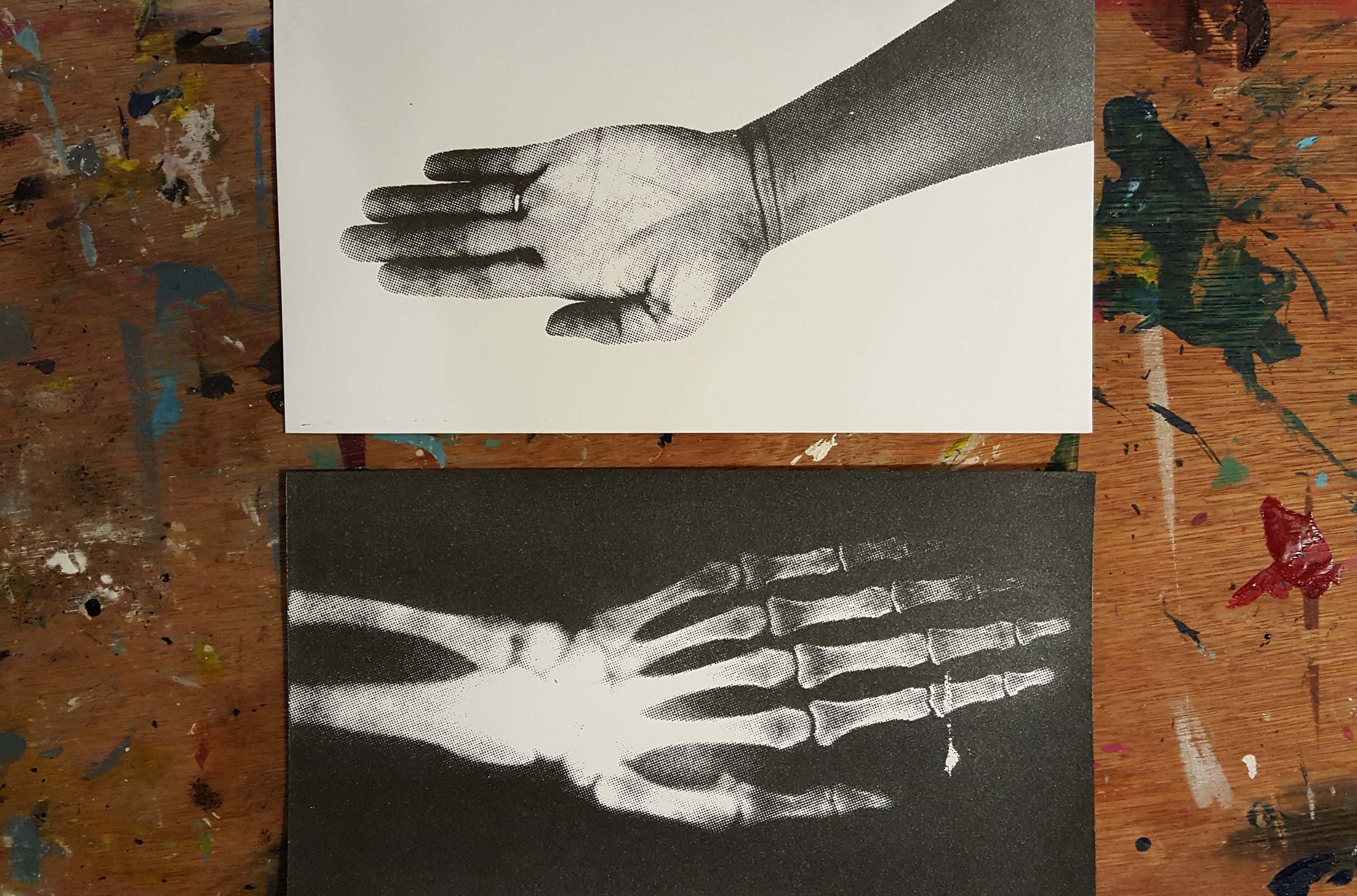 Photograph of an artist's workspace covered in paint, with two art pieces lined up together on it. The top artwork is a silkscreen of a hand, wrist and arm. The second is an x-ray of that same hand, wrist and arm.