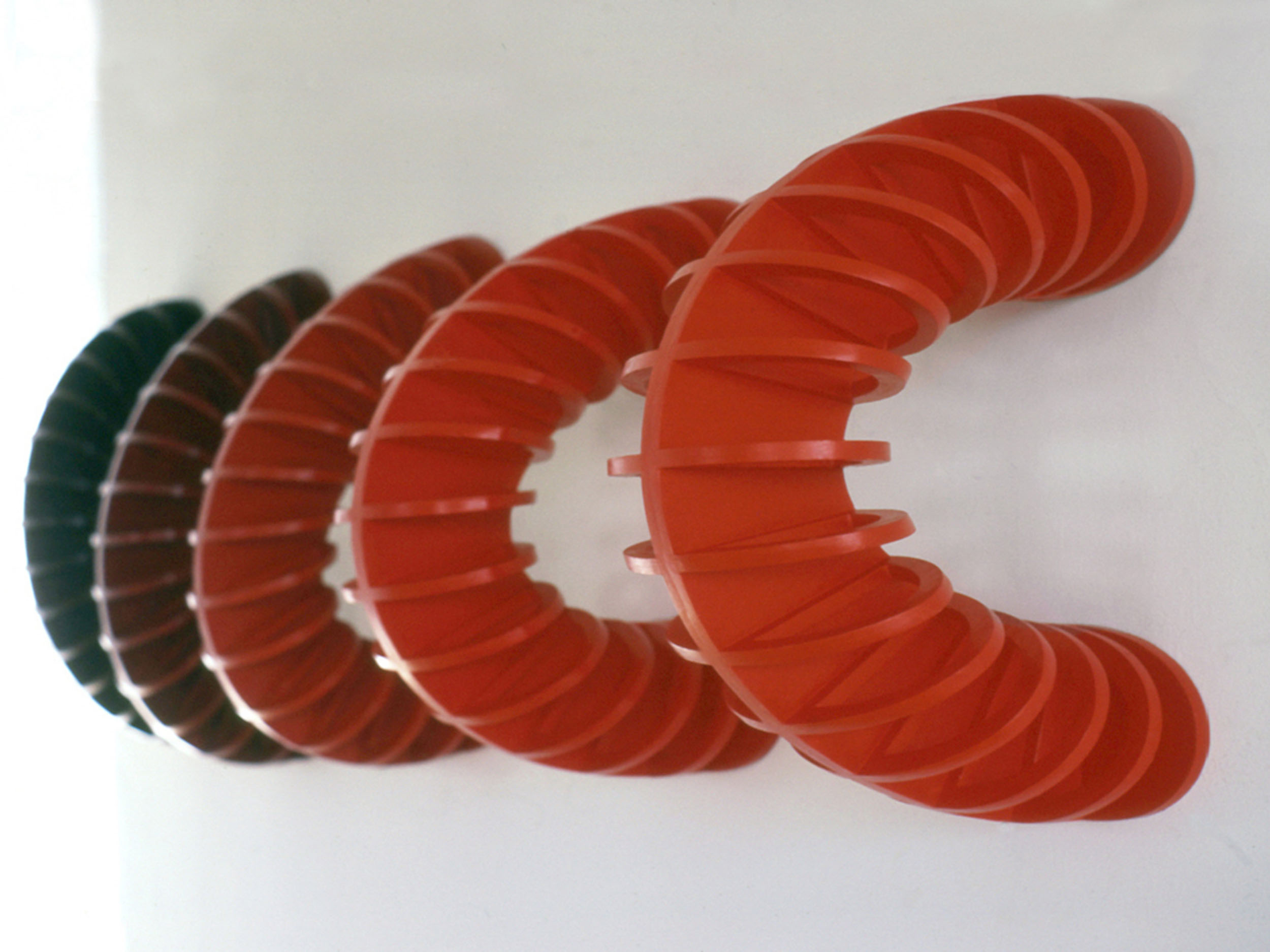 A sculpture comprised of 5 C-shaped semi-circles attached to the wall by their end points in a line. Each is similar to the next except for the color, which is an increasingly darker shade of red for each piece.