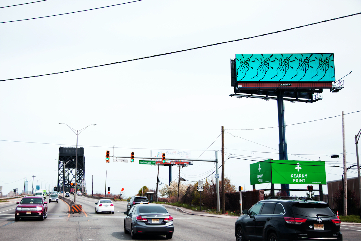 A photograph of a New Jersey roadway that contains a billboard depicting a digital animation of clapping hands.