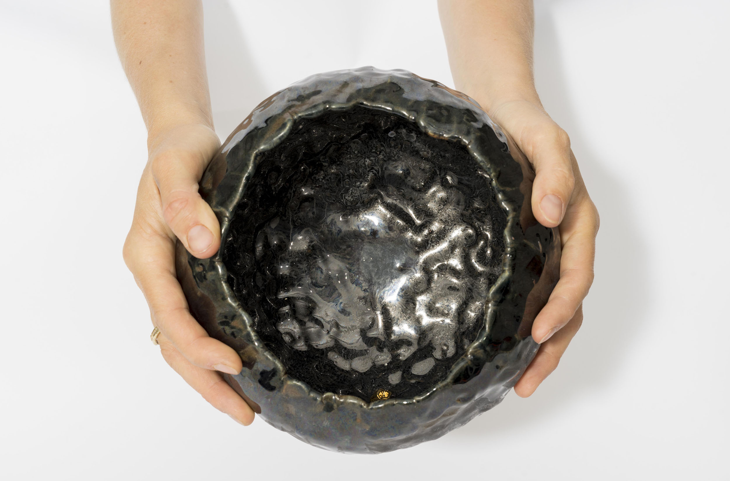 Two hands hold a ceramic art piece that resembles a crater or rough bowl. It is glazed in dark, glossy black and gold.