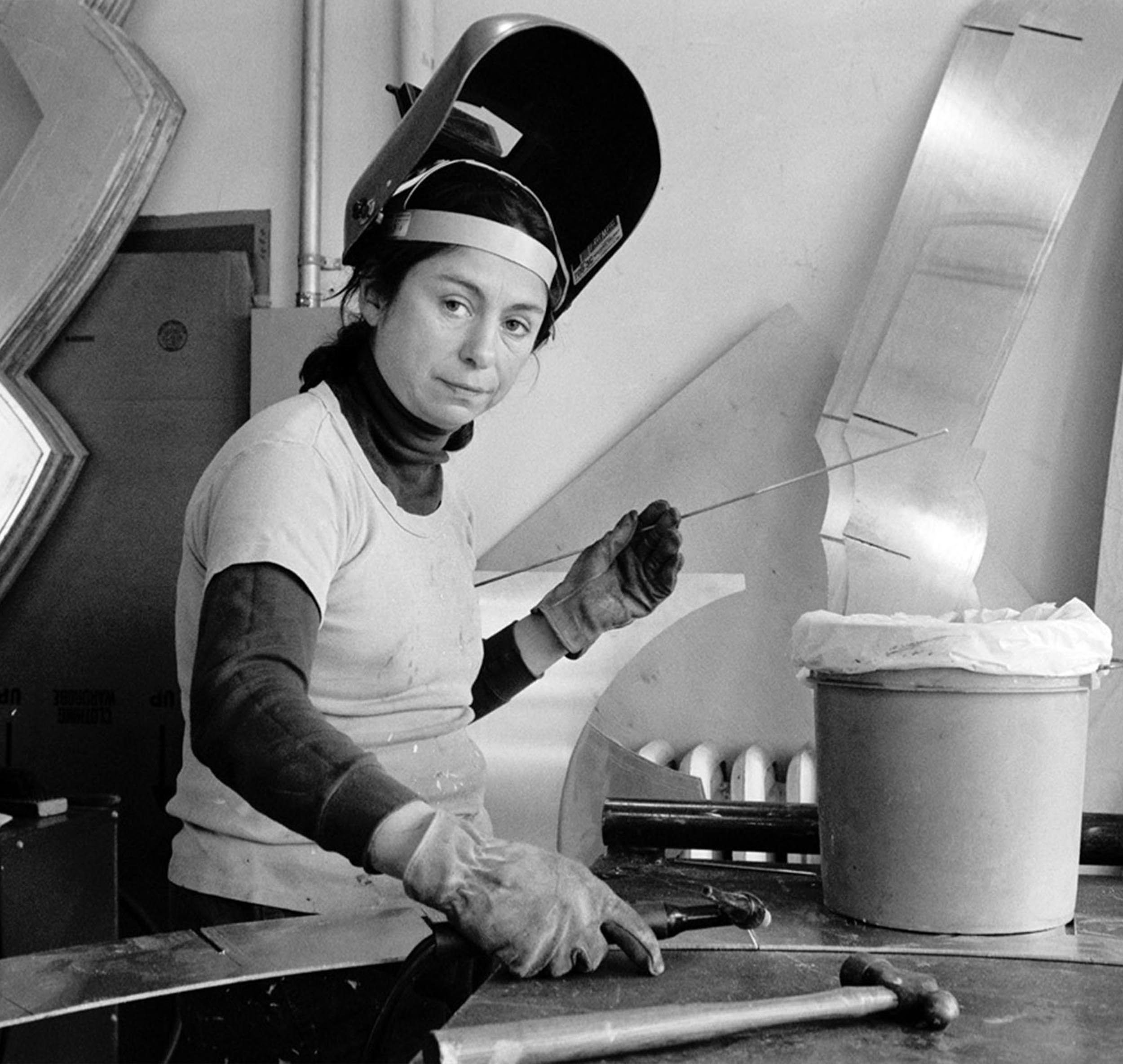 Black and white photograph of Mary Ann Unger at work on her sculpture, wearing work gloves and welder's helmet.