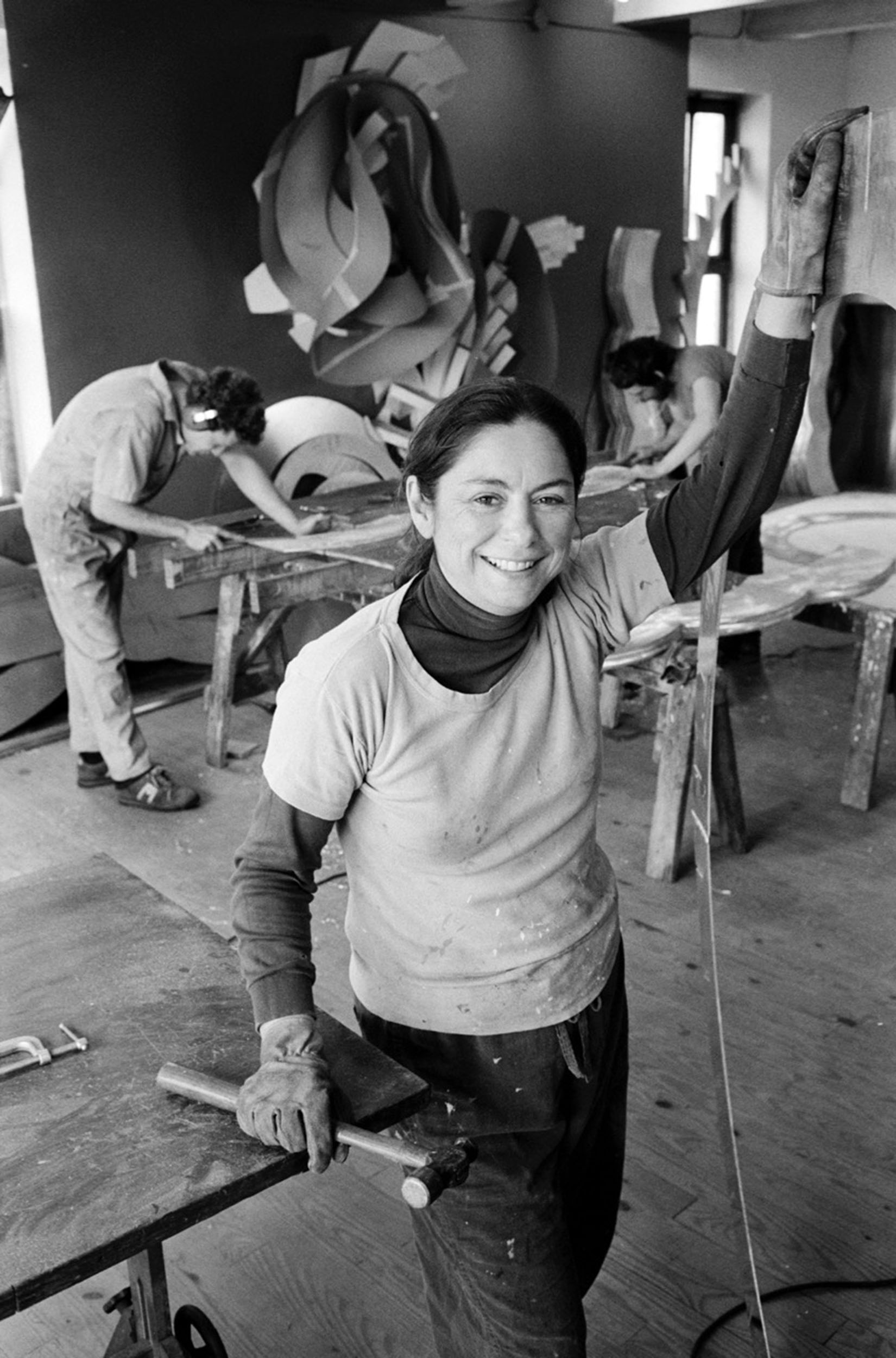 Sculptor Mary Ann Unger at work, wearing work gloves, holding a hammer, and handling metal. She smiles to the camera.