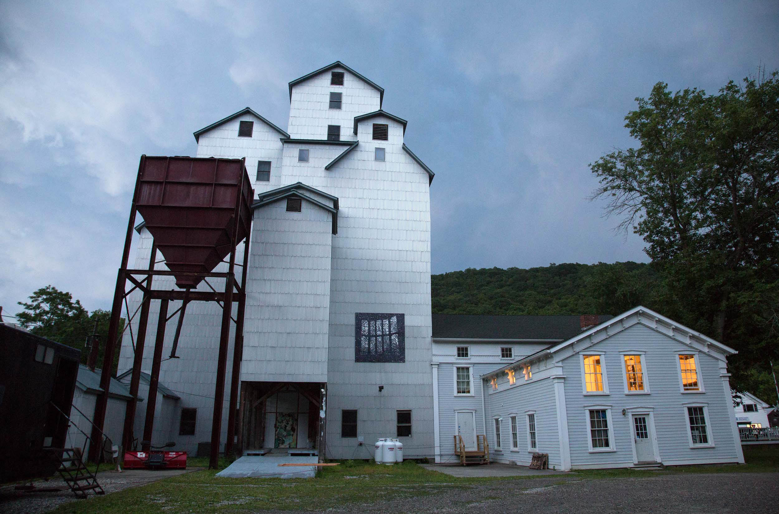 The exterior of the Wassaic Project's home base, called Maxon Mills, under a stormy sky. It is comprised of several interlocking buildings including a former grain elevator and auction barn.