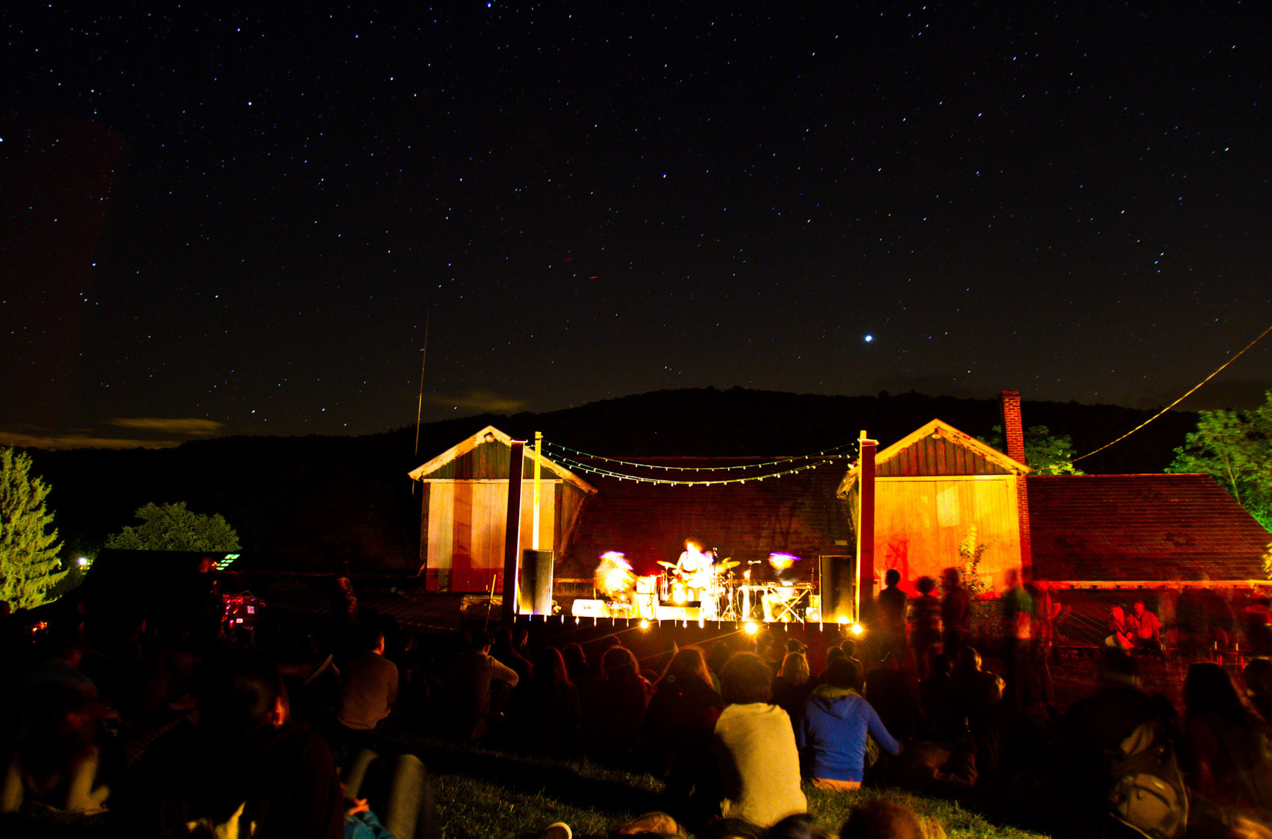 A wide shot photograph depicts an audience watching a band outdoors at the Wassaic Project with a starry sky above them.